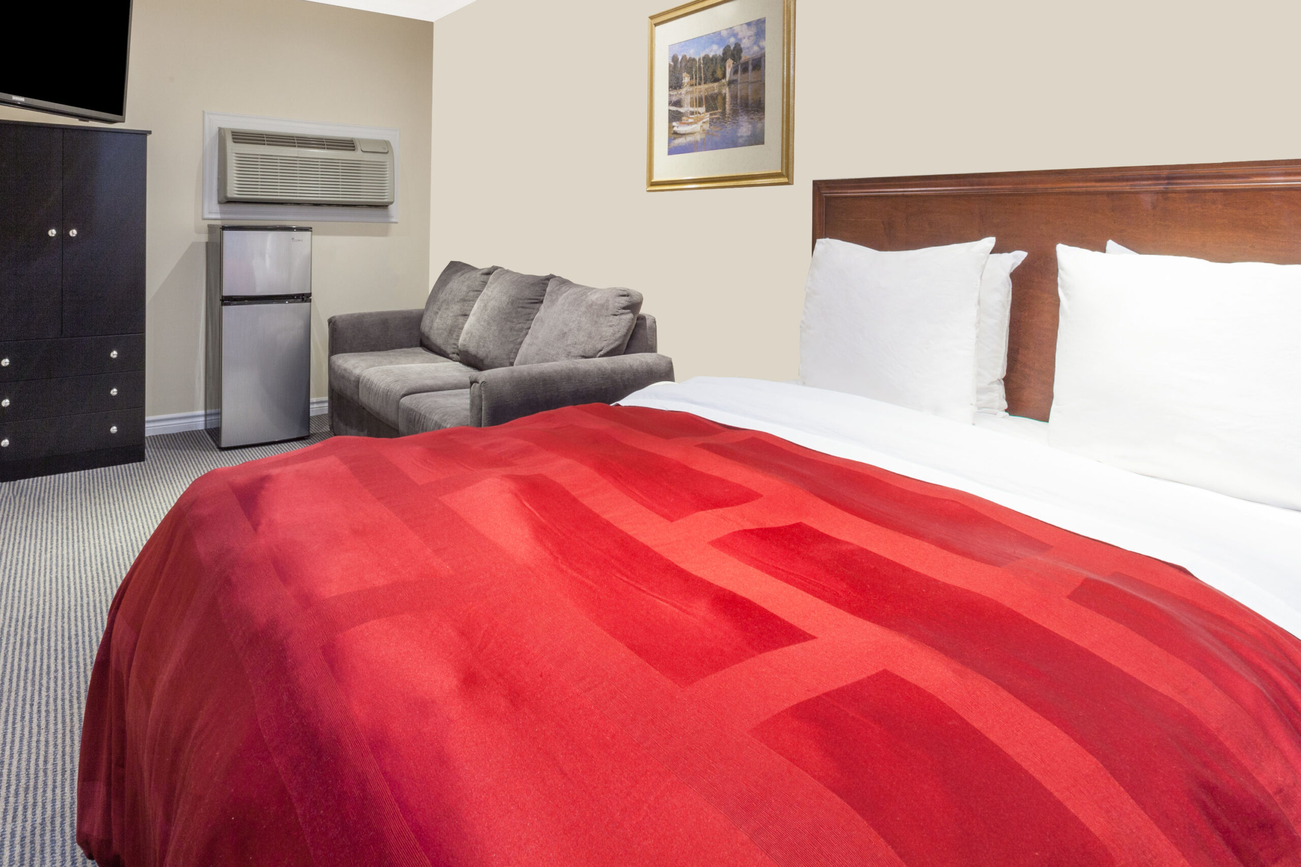 A motel room with a large king-sized bed, two red armchairs by the window, a wooden dresser, and neutral-colored walls.